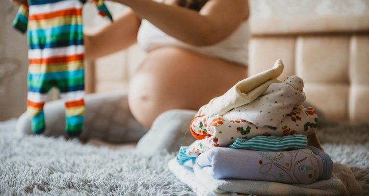 Pile of baby clothes, necessities and pregnant woman on bed in home interior of bedroom. Pregnant woman is getting ready for the maternity hospital, packing baby stuff. Pregnancy, birth concept.
